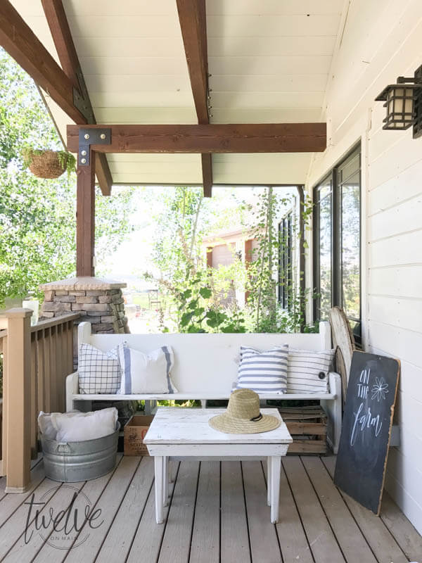 This incredible farmhouse style vintage church pew is the perfect addition to this farmhouse porch! You have to see what it looked like before. With the addition of farmhouse pillows, crates, baskets, and galvanized tubs, this space is perfection.