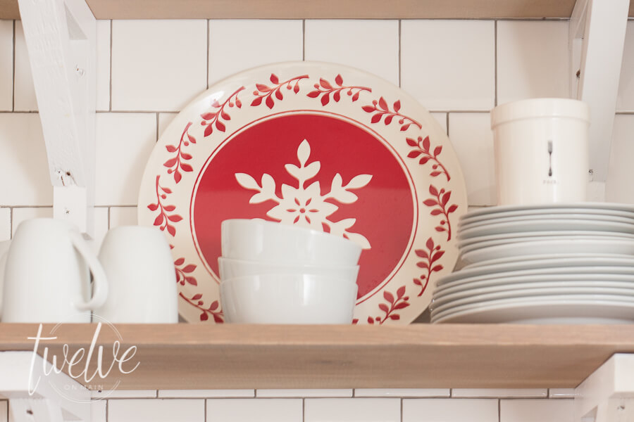 Red and white farmhouse Christmas kitchen decor complete with aRae Dunn Clay pottery, a vintage turkish rug, decorated open shelves, flocked Christmas wreath, and other simple farmhouse Christmas decor pieces.