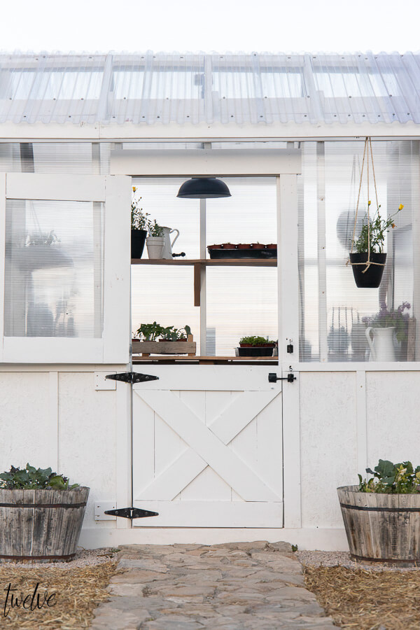 Amazing white DIY greenhouse design complete with a dutch door for ventilation, gorgeous lighting, wood shelves, and so much more. Can you believe this is also super affordable? Check it out!